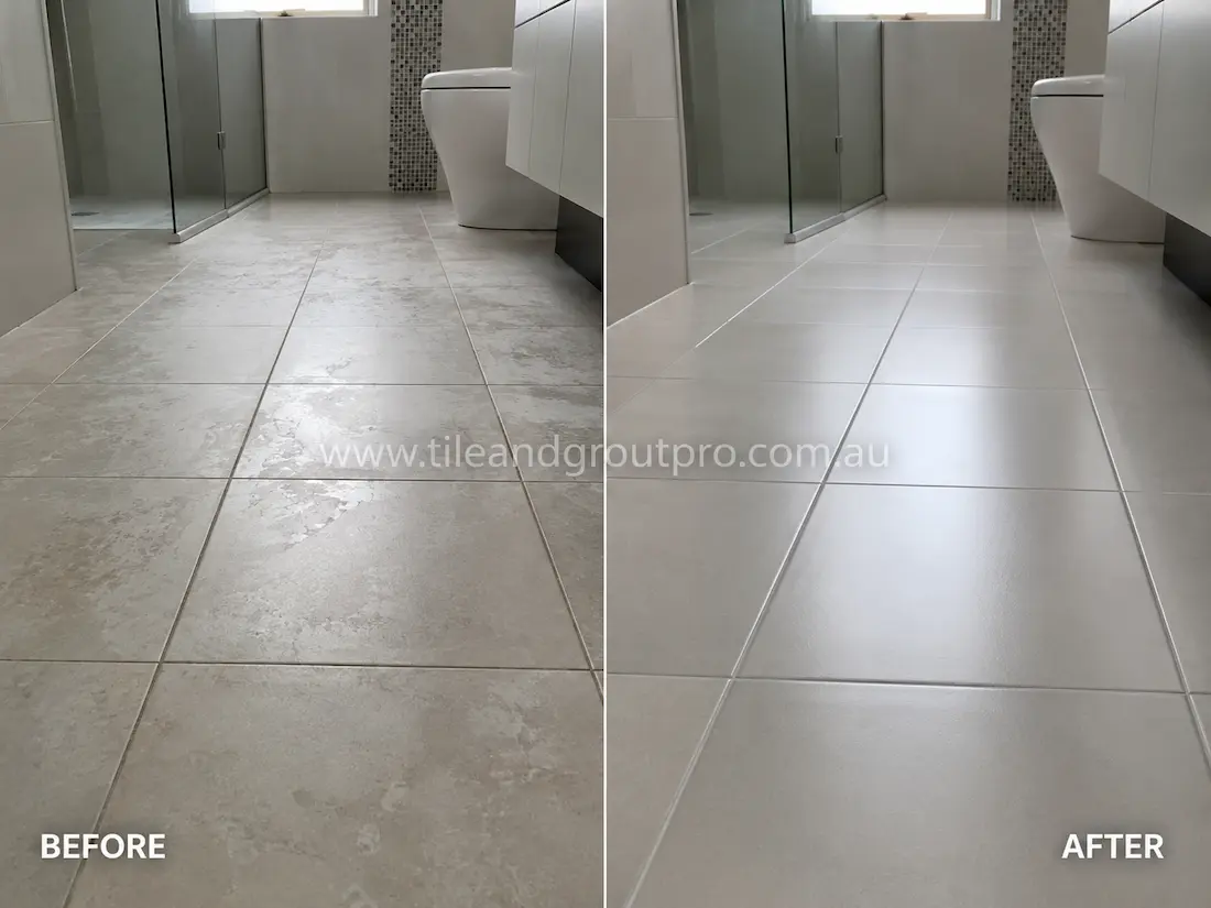 Bathroom floor with light grey ceramic tiles before sealer stripping, showing patchy sealer residue and dull tile surfaces near a glass shower and vanity.