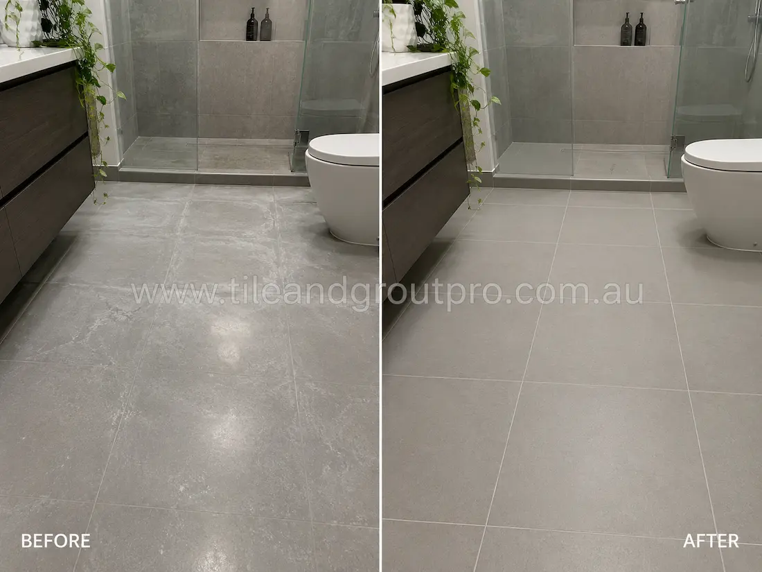 Contemporary bathroom with grey porcelain floor tiles before sealer stripping, featuring visible sealer haze, cloudy patches, and dull tile surfaces near the vanity and glass shower enclosure.